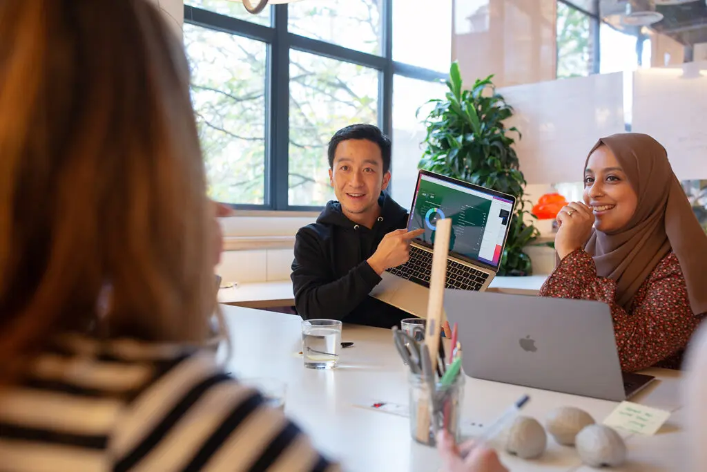 Office meeting scene with one person holding up a laptop displaying a dashboard screen, while others sit around a table engaged and smiling; natural light streams through large windows and a potted plant is visible in the background.