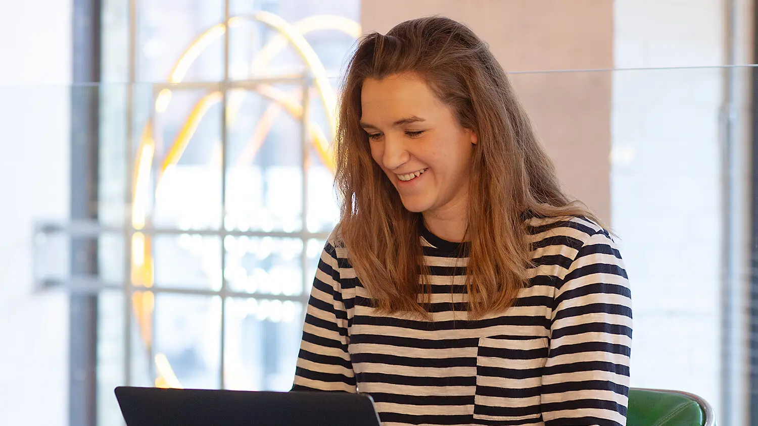 Person smiling while using a laptop, seated indoors in front of a large window with abstract neon lighting in the background; wearing a black-and-white striped top.