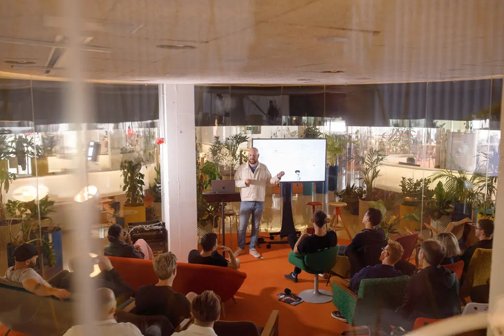 Speaker presenting at a podium with a laptop in a modern indoor setting, backed by plants and a screen displaying a Notion page titled “The Rise of ‘Big Notion Energy’.” An audience member is visible in the foreground.