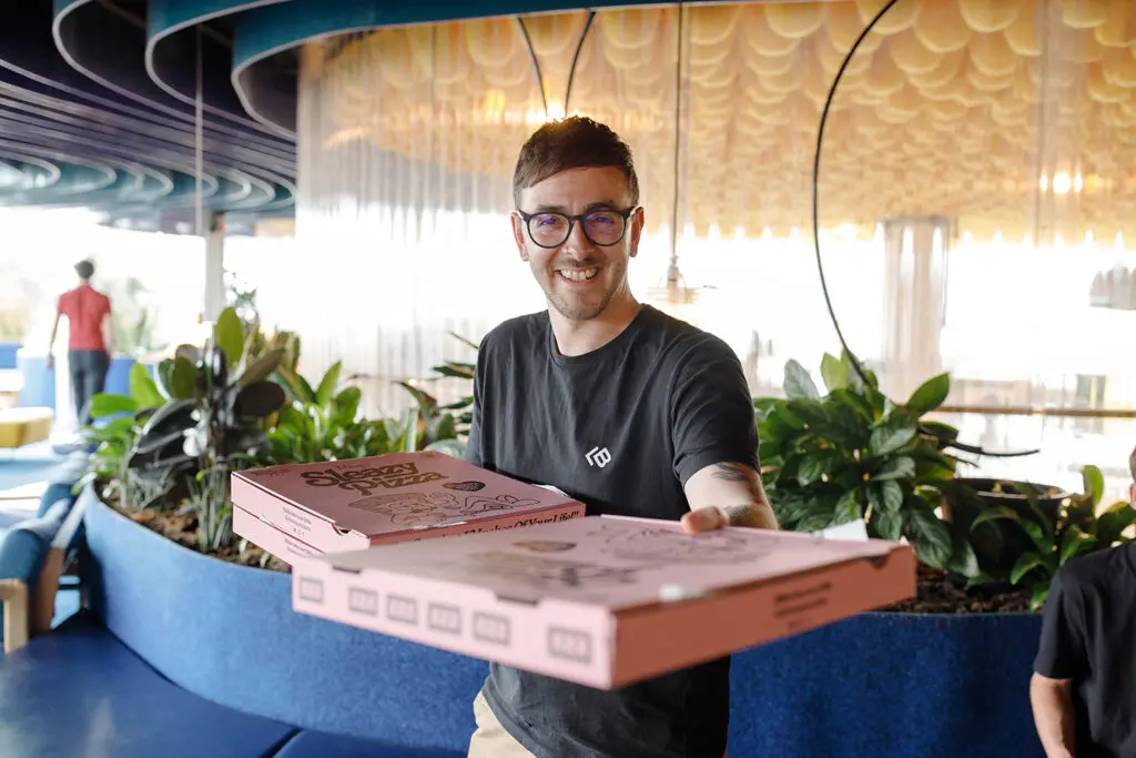 Smiling person in a black T-shirt offering two pink pizza boxes toward the camera, standing in a modern, plant-filled indoor space with curved blue planters and a warm yellow ceiling installation.