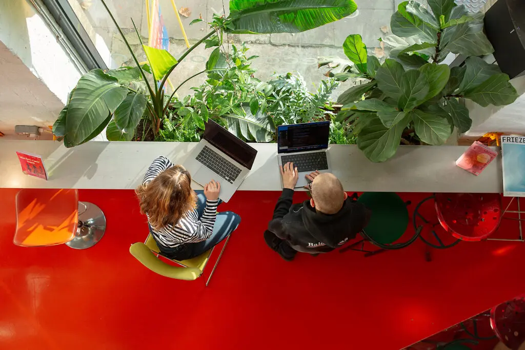 Overhead view of two people working on laptops at a counter beside large leafy plants and a tall window, with bright red flooring and mismatched chairs creating a vibrant, creative workspace atmosphere.