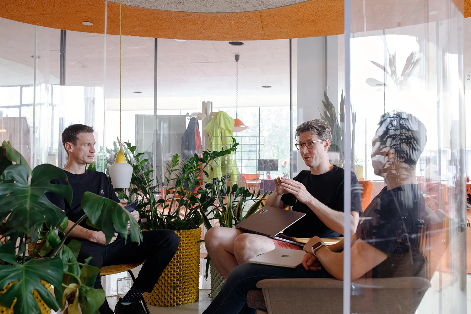 Three InfoSec leaders having a discussion in a glass-walled meeting pod surrounded by lush indoor plants, with laptops open and one person gesturing while speaking.