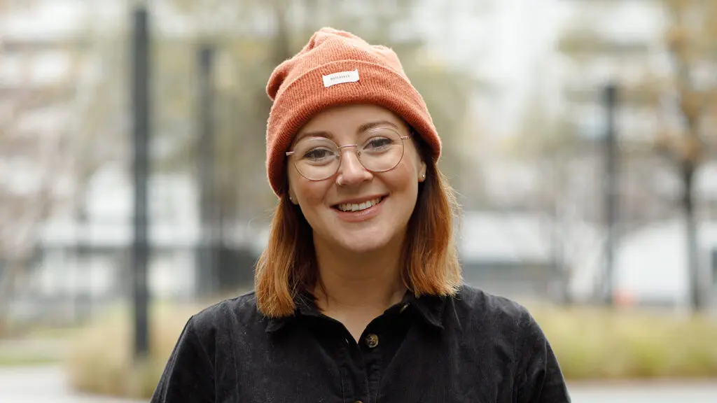 Person smiling outdoors with hands in pockets, wearing a burnt orange beanie, glasses, and a black button-up jumpsuit, with a blurred cityscape in the background.