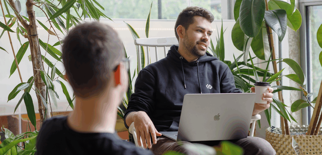 LeftBrain team member chats with a colleague in a plant-filled office, holding coffee and working on a laptop.
