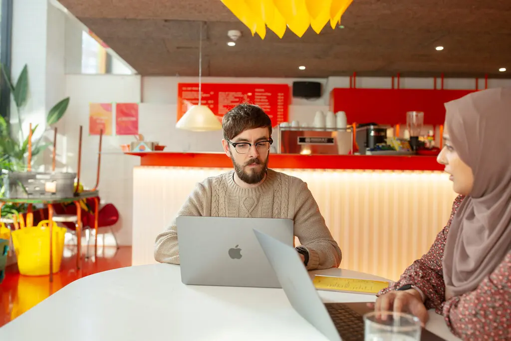 Two people sitting at a white table with laptops in a colourful café-style workspace, engaged in conversation. A bright red counter, warm lighting, and plants create a vibrant, relaxed atmosphere.