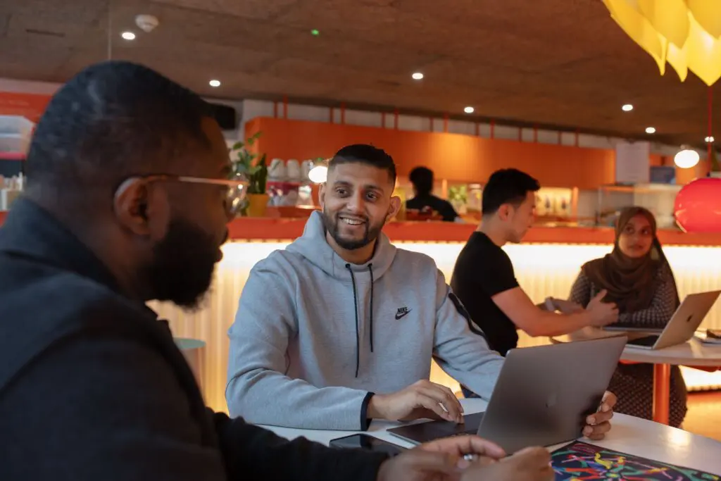 Two people in the foreground chatting and working on laptops in a modern, warmly lit café-style workspace; in the background, another pair sits at a high table also engaged in conversation with laptops open.
