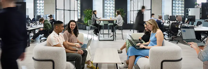 Open-plan office space with people working and chatting in small groups; some sit on modern lounge chairs with laptops, while others work at desks or stand around a high table. The environment is bright and contemporary with large windows, plants, and a clean, minimalist design.