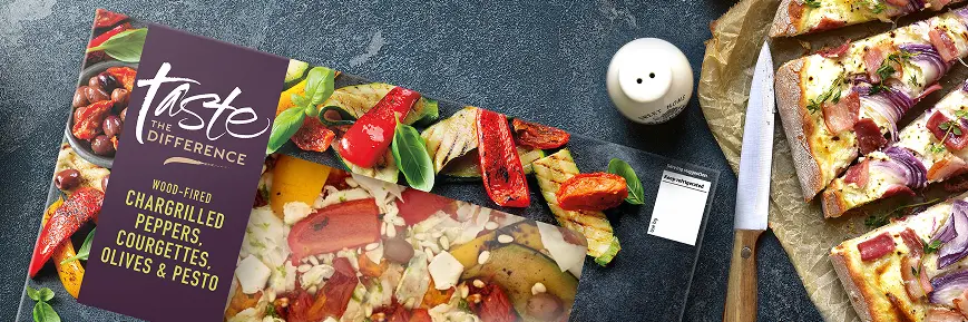 The image shows a boxed supermarket pizza from the "Taste the Difference" range. The flavour is “Wood-Fired Chargrilled Peppers, Courgettes, Olives & Pesto” on a hand-stretched sourdough base. The pizza box is placed on a dark stone surface alongside a sliced cooked pizza topped with red onion and bacon, a knife, and a set of salt and pepper shakers.