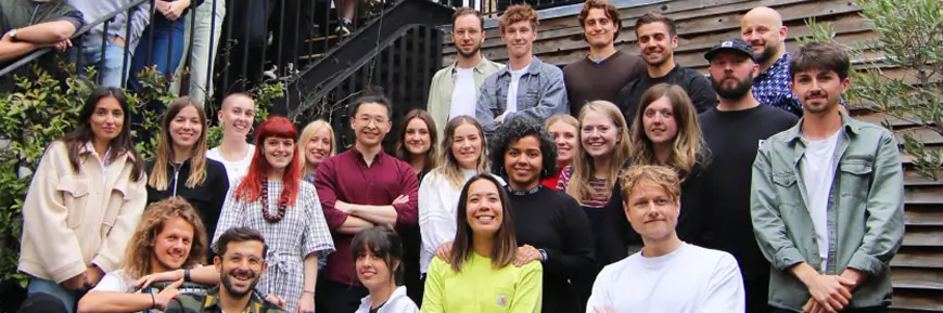 A group photo of around 30 people standing and sitting on outdoor stairs and in front of a wooden wall, smiling and posing together in casual clothing. Shrubs and greenery frame the sides of the image.