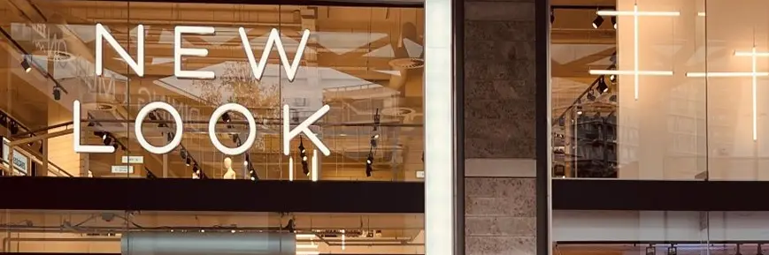 Storefront with large glass windows featuring the bold text “NEW LOOK” in white capital letters; the interior shows modern lighting, mannequins, and retail displays.