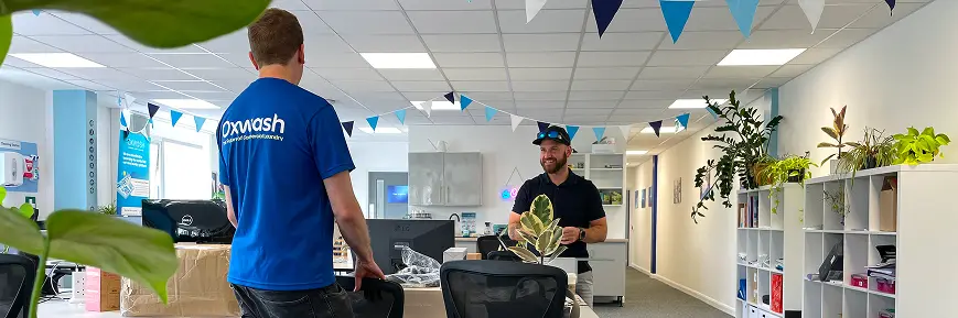 Two people talking in a brightly lit office decorated with blue and white bunting, surrounded by plants, desks, and shelving. One person wears an “Oxwash” T-shirt.