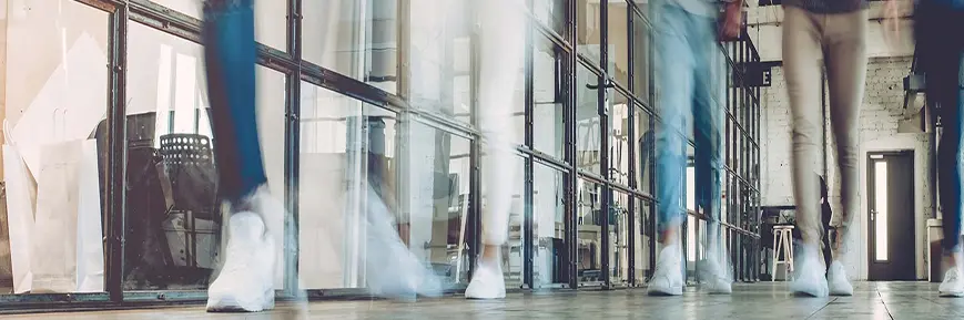 Blurred motion image of people walking through a modern office hallway with glass walls, exposed brick, and visible shopping bags and office furniture.