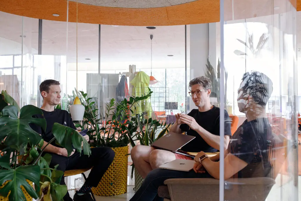 Three people in discussion inside a glass meeting pod surrounded by lush green plants, with laptops in hand in a bright, modern office.