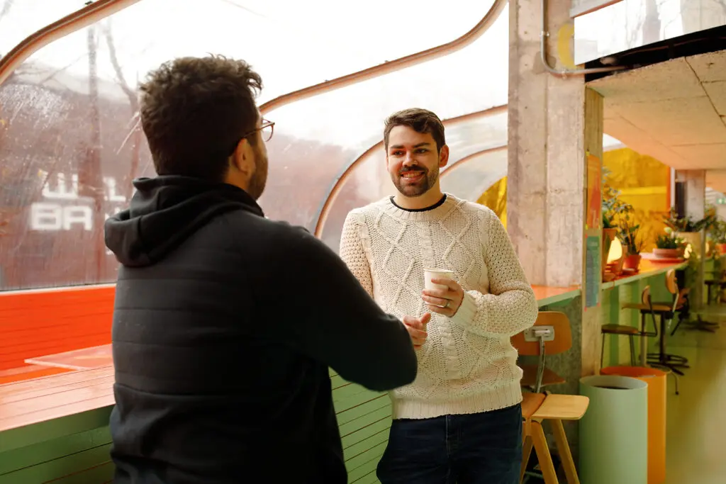 Two people chatting indoors by a curved window, one holding a coffee cup and smiling, surrounded by warm-toned décor and potted plants.