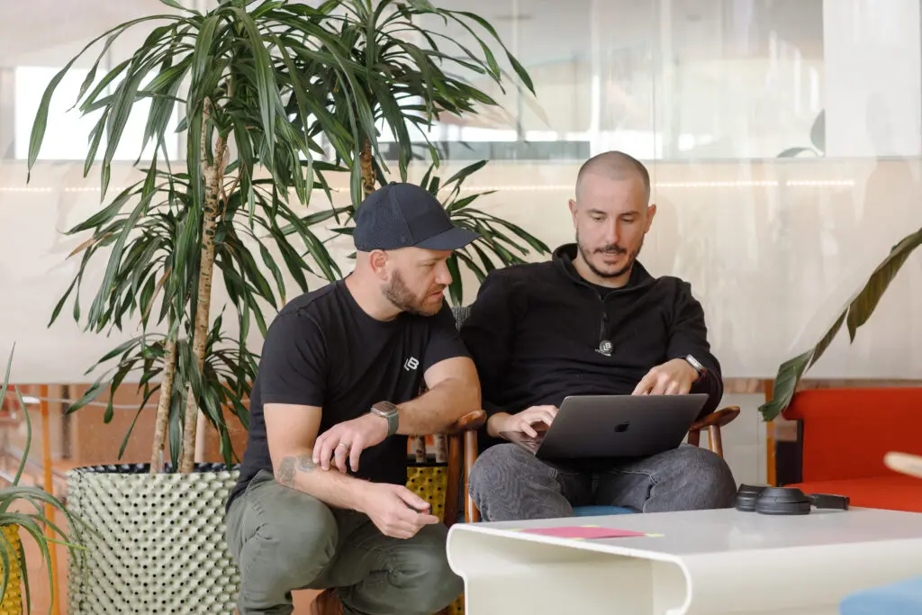 Two people sitting closely together in a relaxed office setting, discussing something on a laptop screen. One is seated while the other kneels beside, with large plants and a white table with headphones and notes in the foreground.