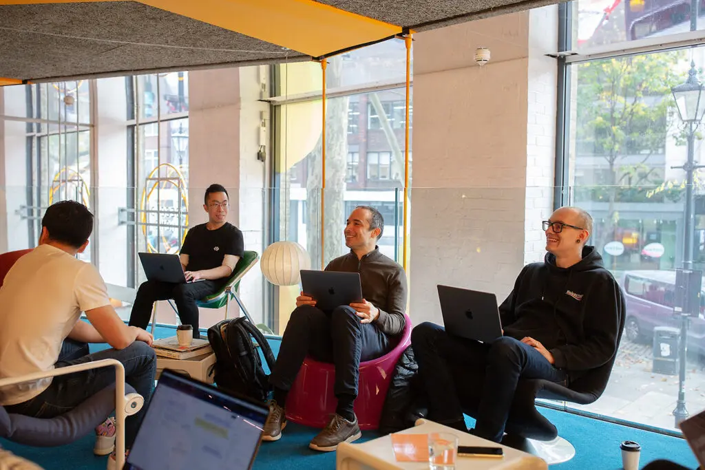 Group of people sitting in a modern office lounge, each with a laptop, engaged in conversation near large windows overlooking a city street.