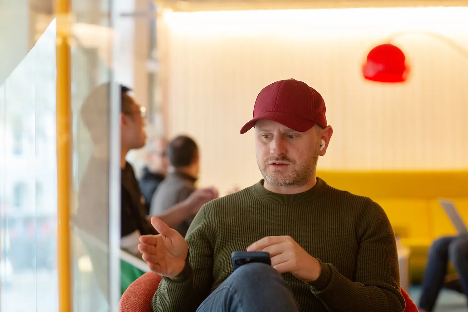 Person in a red cap and green jumper sitting in a modern lounge chair, gesturing while looking at a phone, with others working in the background.