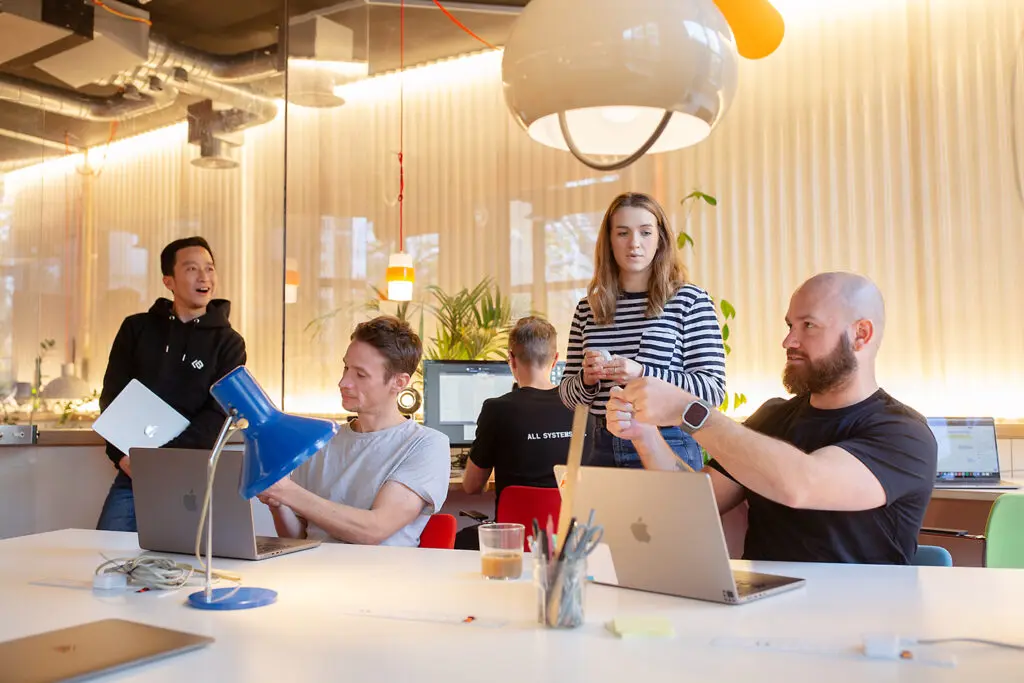Group of five people in a modern office setting, interacting casually around laptops and a table with a blue desk lamp, wires, and drinks. One person is adjusting a cable, another is standing with a drink, and a person at the back is working on a monitor.