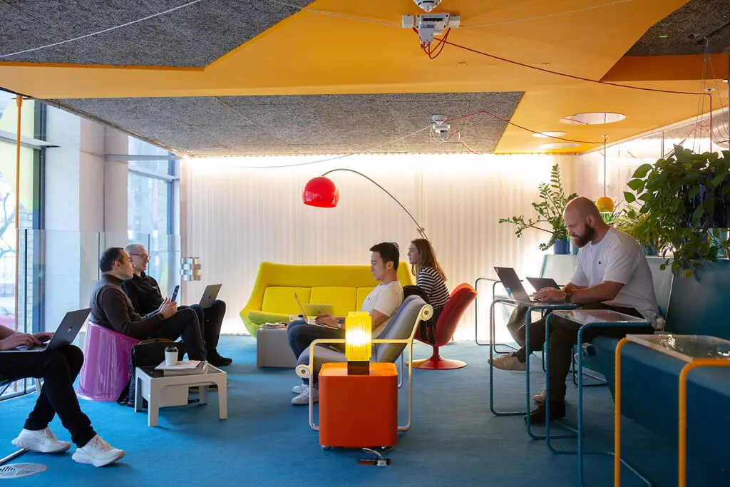 Six people working on laptops in a vibrant, lounge-style office space with colourful furniture, hanging lights, and lush plants. The setting includes a yellow ceiling, red lamp, and a bright yellow couch, creating a relaxed and creative atmosphere.