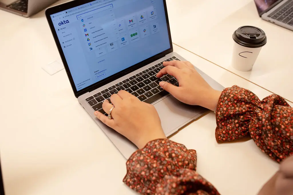 Close-up of hands typing on a laptop displaying the Okta login dashboard, with a coffee cup and other laptops visible on a white desk.