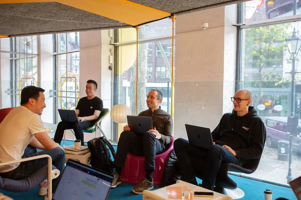 Four people having a relaxed conversation in a brightly lit modern office lounge with large street-facing windows; three are seated with laptops, and one is leaning forward with a smile. A coffee cup, backpack, and lamp are visible, adding to the casual, collaborative setting.