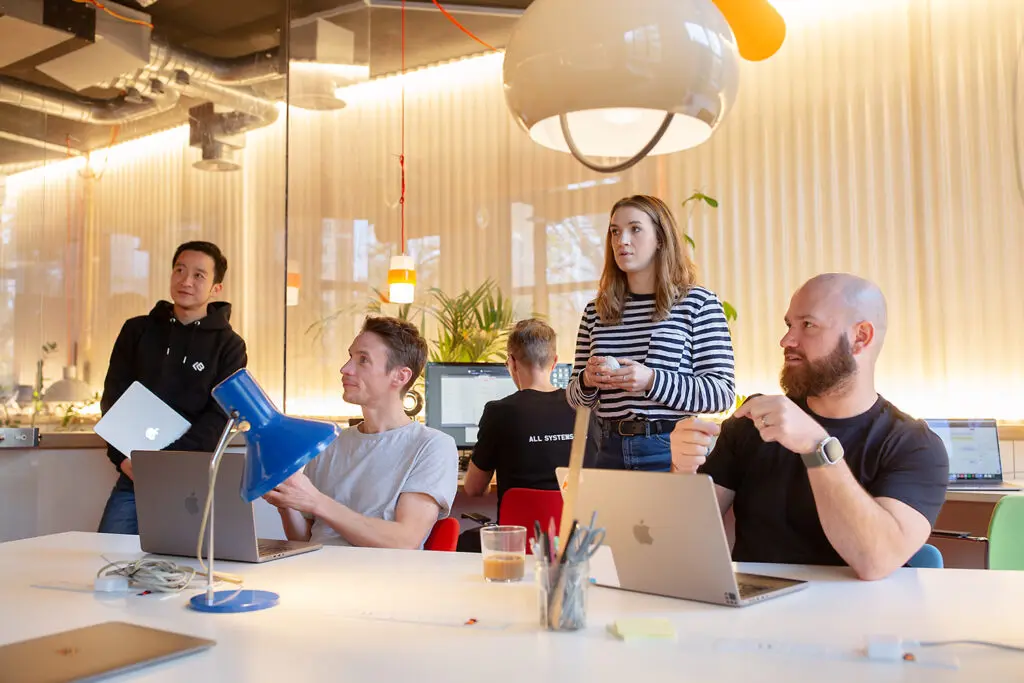 A group of six people in a bright, modern office engaged in discussion; some are seated at a table with laptops, while others stand nearby. The setting includes warm lighting, indoor plants, and a large overhead lamp, creating a collaborative and informal workspace atmosphere.
