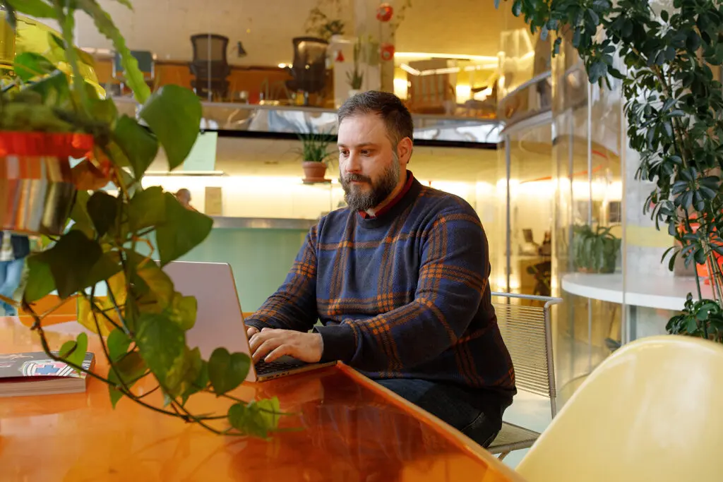 Person seated at a bright orange desk, typing on a laptop in a stylish, plant-filled office space with warm lighting and glass walls. A patterned jumper and nearby greenery add texture and colour to the relaxed environment.