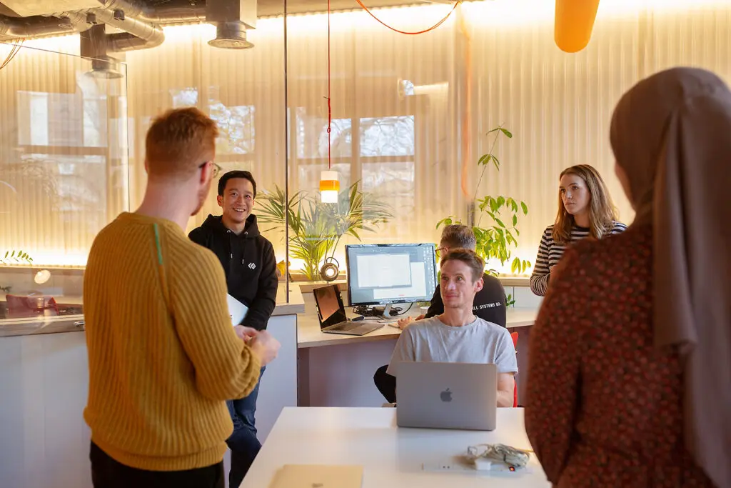 Group of six people in a warmly lit office space, some standing and some seated around desks with laptops and monitors, engaged in a relaxed team discussion. Indoor plants and ambient lighting add to the comfortable atmosphere.