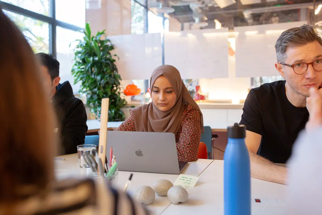 A group of people seated around a bright meeting table in a modern office; one person in the centre is focused on a laptop, with others listening or thinking. The table holds a water bottle, pens, stress balls, and sticky notes, with whiteboards and plants in the background.