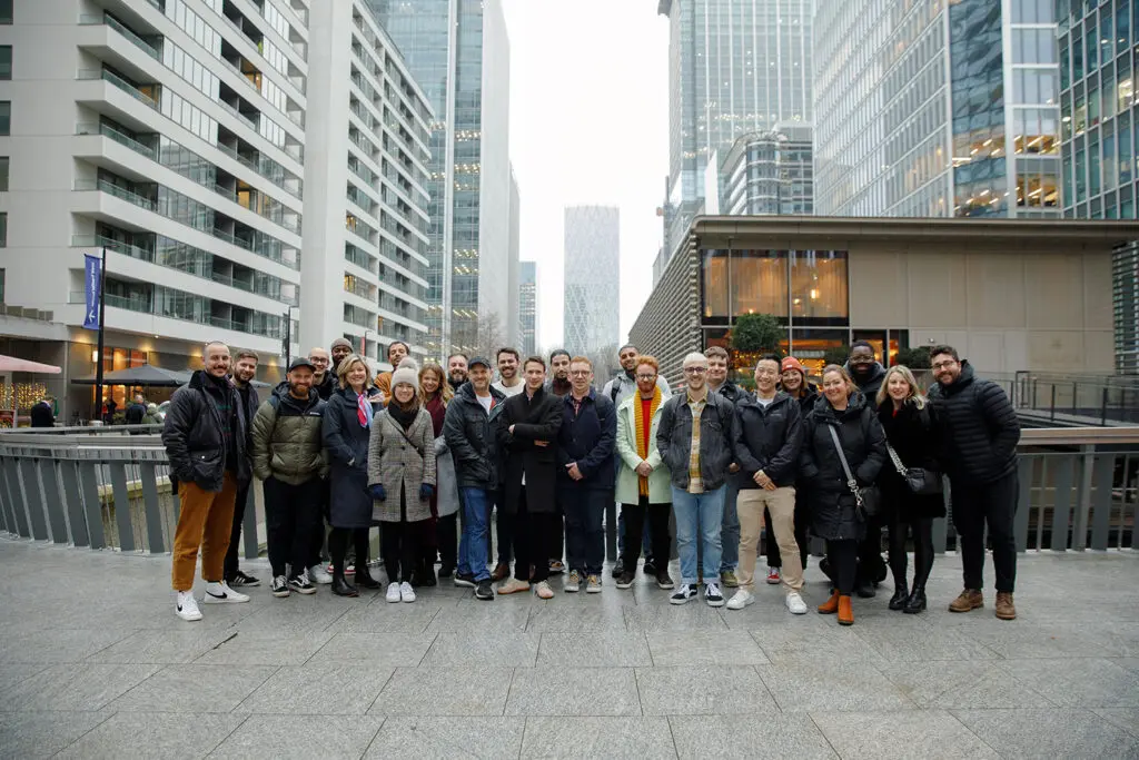 Group of people standing and smiling for a team photo in an urban setting, surrounded by tall modern buildings and overcast skies.