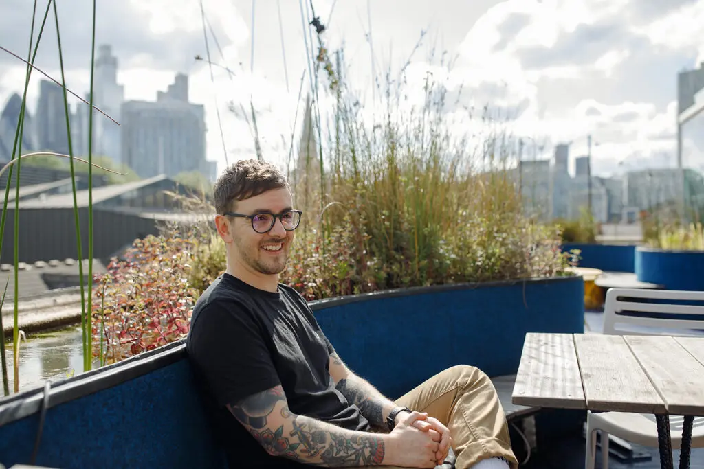 Person with short hair, glasses, and tattooed arms sits smiling on an outdoor bench surrounded by tall grasses and plants, with a city skyline in the background.