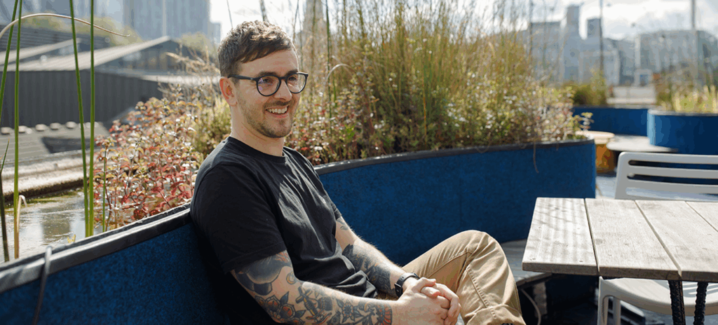 Person with short hair, glasses, and tattooed arms sits smiling on an outdoor bench surrounded by tall grasses and plants, with a city skyline in the background.