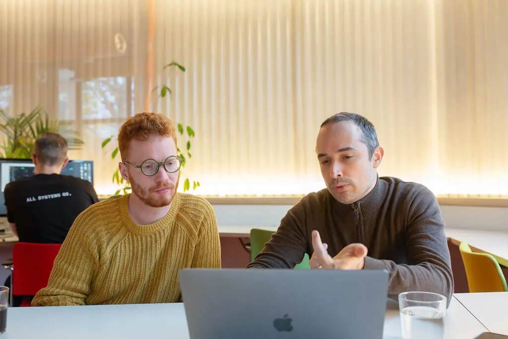 Two people sitting at a table, focused on a laptop screen, with one gesturing as if explaining something. Another person works at a computer in the background, wearing a shirt that reads “ALL SYSTEMS GO.”