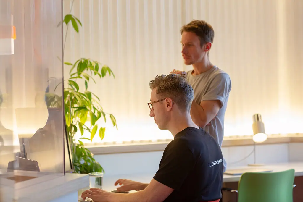 Two people working together at a desk in a brightly lit office. One is seated and typing on a keyboard while the other stands behind, watching attentively. A leafy plant, glass of water, and desk lamp are visible in the background.