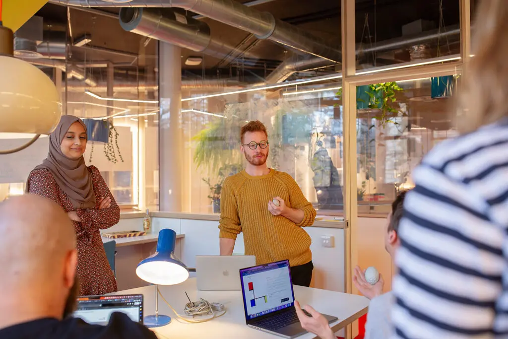 Five people gathered around a desk in a bright, modern office space, with laptops open and two individuals holding stress balls, engaged in a casual team discussion.