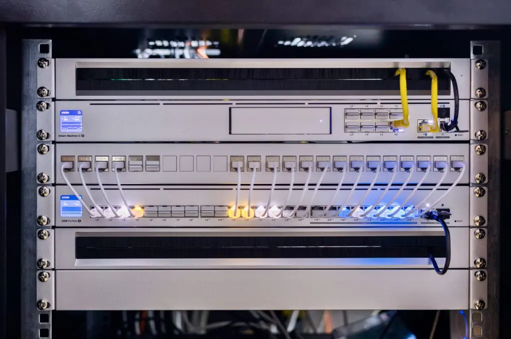 Close-up of a sleek, silver server rack with multiple network cables plugged into ports, LED indicators lit in blue and orange, and a “Dream Machine” and “USW Pro Max” label visible.
