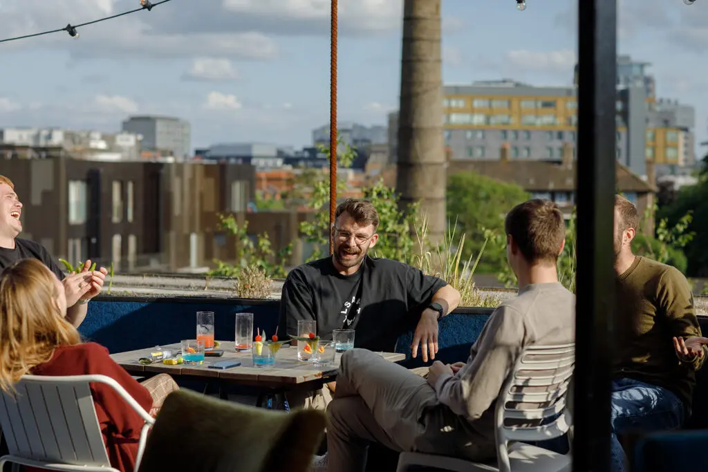 Group of people sitting around a table on a rooftop terrace, laughing and chatting in the sunshine; colourful drinks and garnishes are scattered on the table, with city buildings in the background.