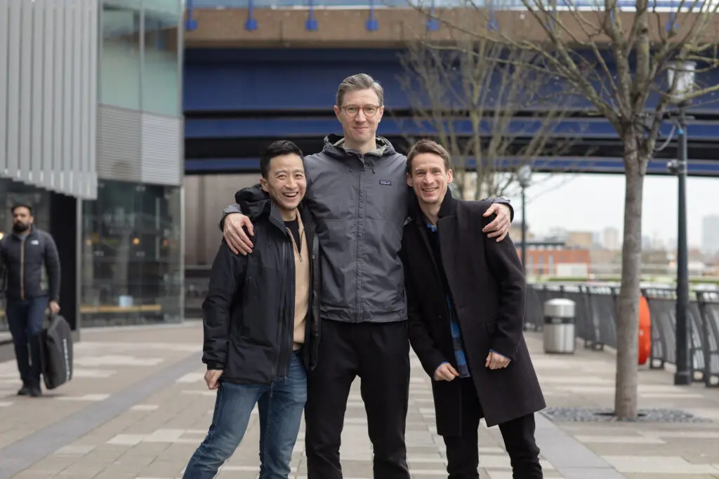 Three people standing outdoors with arms around each other, smiling in front of a modern urban backdrop with bare trees and buildings in the distance.