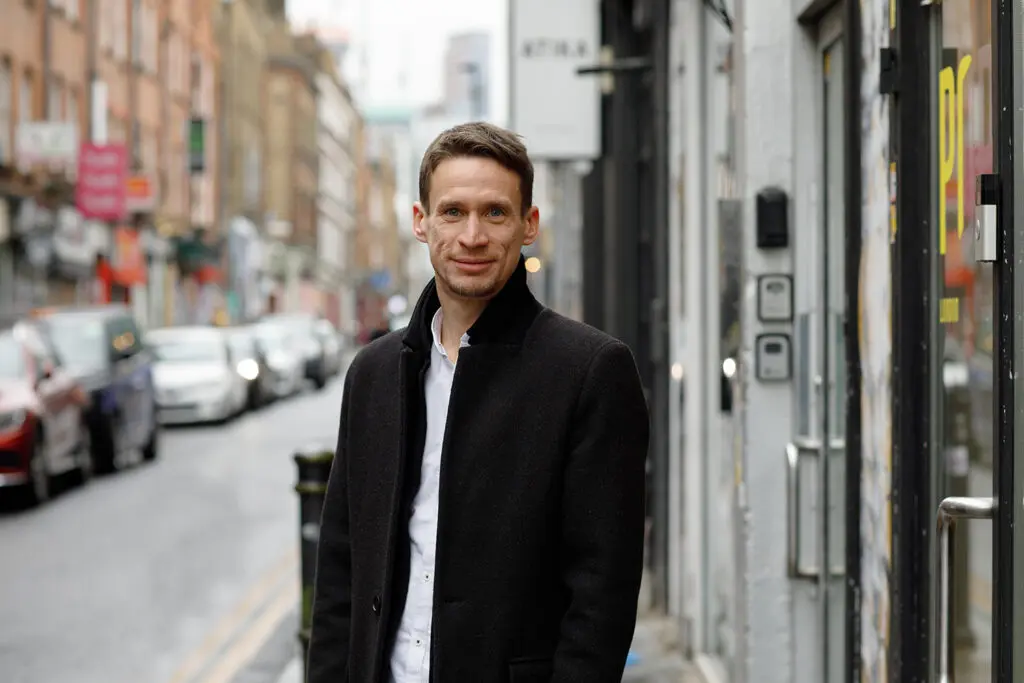 Person standing on a city street in a dark coat and white shirt, with parked cars and blurred shopfronts lining the background.