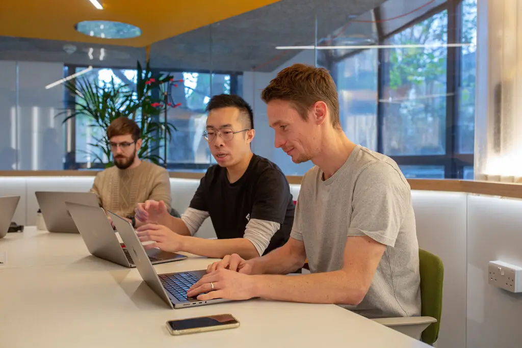 Three people sitting at a white desk working on laptops in a modern office with large windows and indoor plants; one is speaking while the others type. Learn how VC-backed startups and scaleups can lay strong foundations for secure, scalable growth. Discover common pitfalls, practical systems, and why security clarity matters from day one.