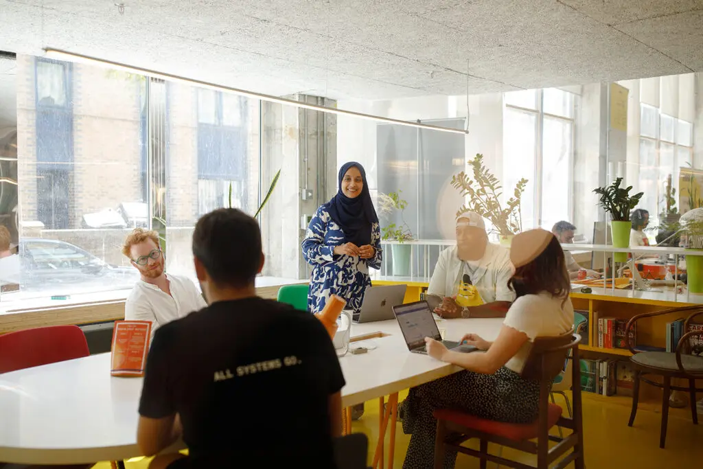 A group of five people gathered around a table in a bright, modern co-working space. One person stands and speaks while the others sit listening or working on laptops. Large windows, indoor plants, and colourful furniture create a relaxed, collaborative atmosphere.