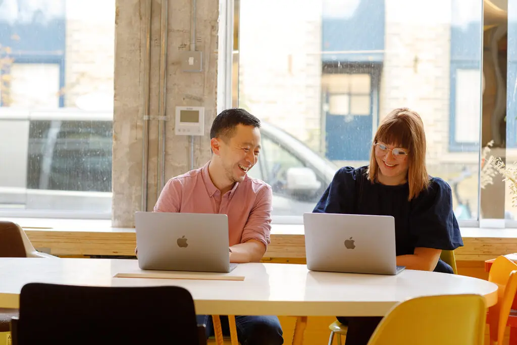Two colleagues sit at a round table with open laptops, smiling and chatting in a bright office space with large windows and colourful chairs.