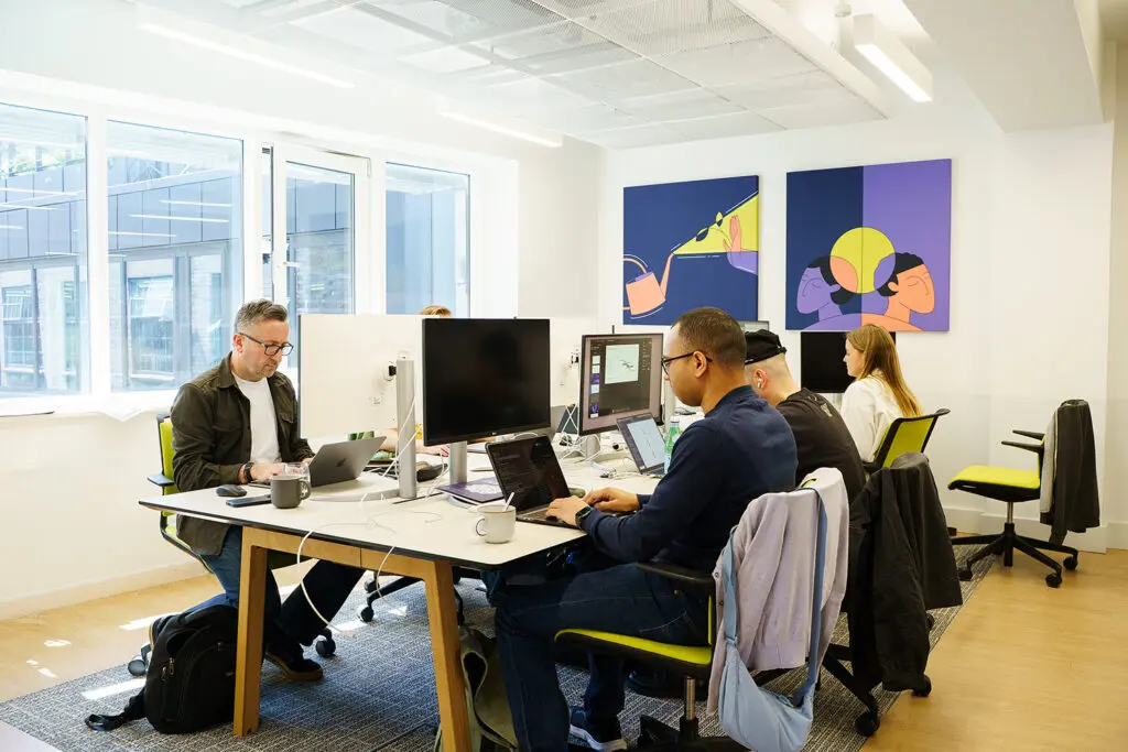 Five people working at shared desks in a bright open-plan office, with laptops and monitors. Two bold illustrated artworks in navy, purple, and yellow hang on the wall behind them.
