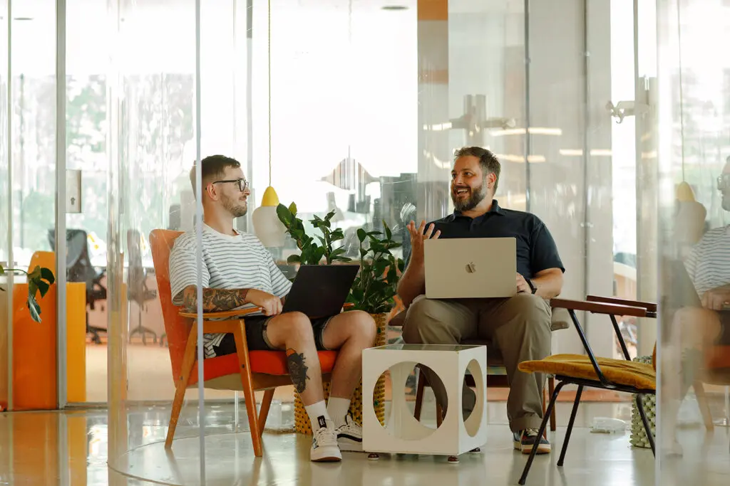 Two colleagues chatting in a modern office lounge, each with a laptop open on their lap — one seated in an orange chair, the other in a matching armchair, separated by a glass-and-white side table, with glass partition walls and a hanging pendant light in the background.