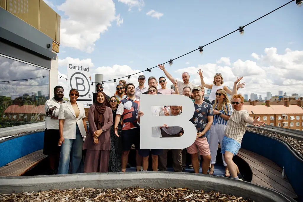 A group of about twenty colleagues celebrating on a rooftop terrace, gathered around a large white letter "B" and a "Certified B Corporation" sign. City rooftops and a partly cloudy sky are visible behind them, with string lights strung overhead. Several people have their arms raised in celebration, and two people are peeking through the holes in the "B" sculpture.