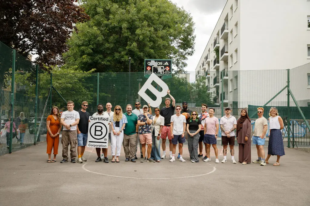 A group of colleagues standing in a line on an outdoor basketball court, posing for a photo with a "Certified B Corporation" sign and a large white letter "B" held overhead. Green fencing, trees, and a residential building are visible in the background, with one person holding a basketball.