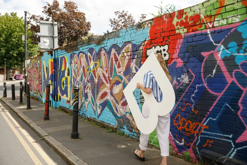 A person in a striped shirt and white trousers carries a large white letter "B" while walking along a sidewalk beside a wall covered in colourful graffiti murals. Trees and a residential street are visible in the background.