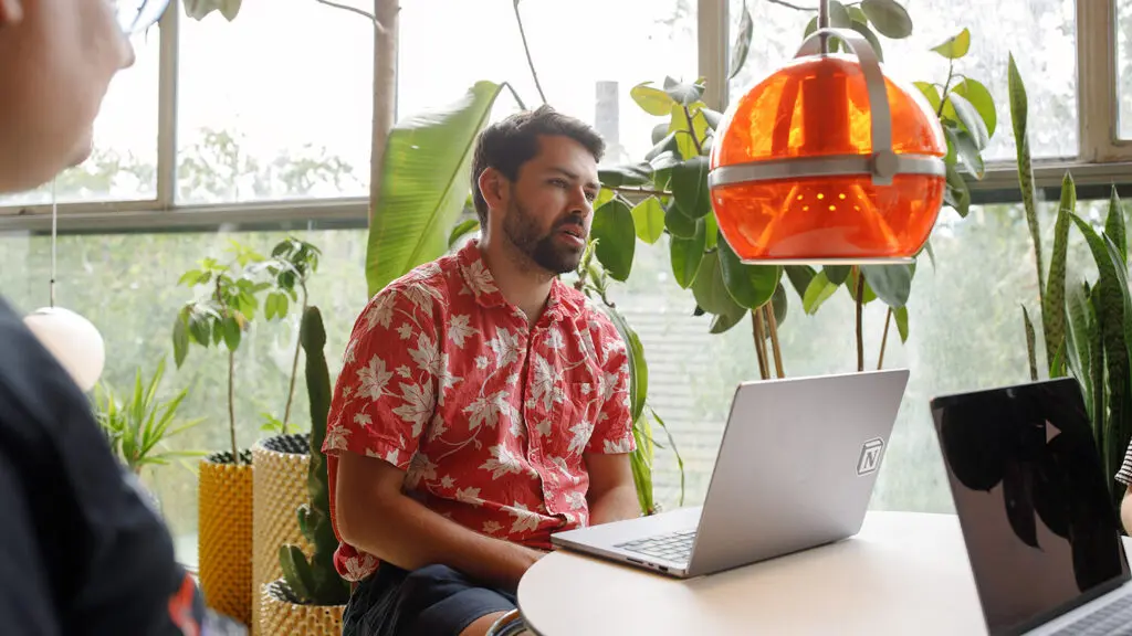 A person with short dark hair and a beard, in a red shirt with a white leaf print, sits talking at a round white table with an open laptop. An orange globe pendant light hangs beside them, and large windows behind them are lined with leafy plants. In the foreground, a second person in glasses is out of focus, facing them.