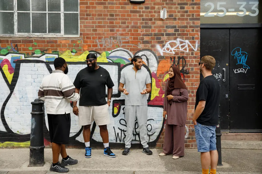Five people stand chatting and laughing on a pavement in front of a brick wall covered in colourful graffiti. One, in a striped top, has their back to the camera. Next to them is someone in a black T-shirt, sunglasses and a durag, then someone in a grey zip-neck top and joggers who is gesturing mid-story. Beside them, a person in a mauve hijab and matching outfit is smiling. On the right, someone in a black T-shirt and denim shorts stands by a black door marked "25–33