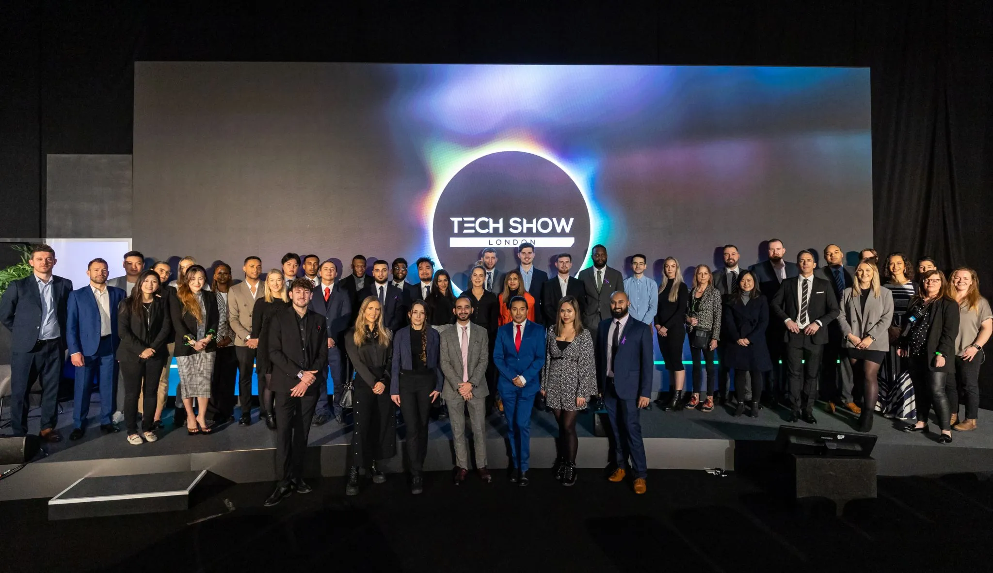 Large group of people in business attire standing on stage in front of a screen displaying “TECH SHOW LONDON” with a dark circular logo and colourful light effects.
